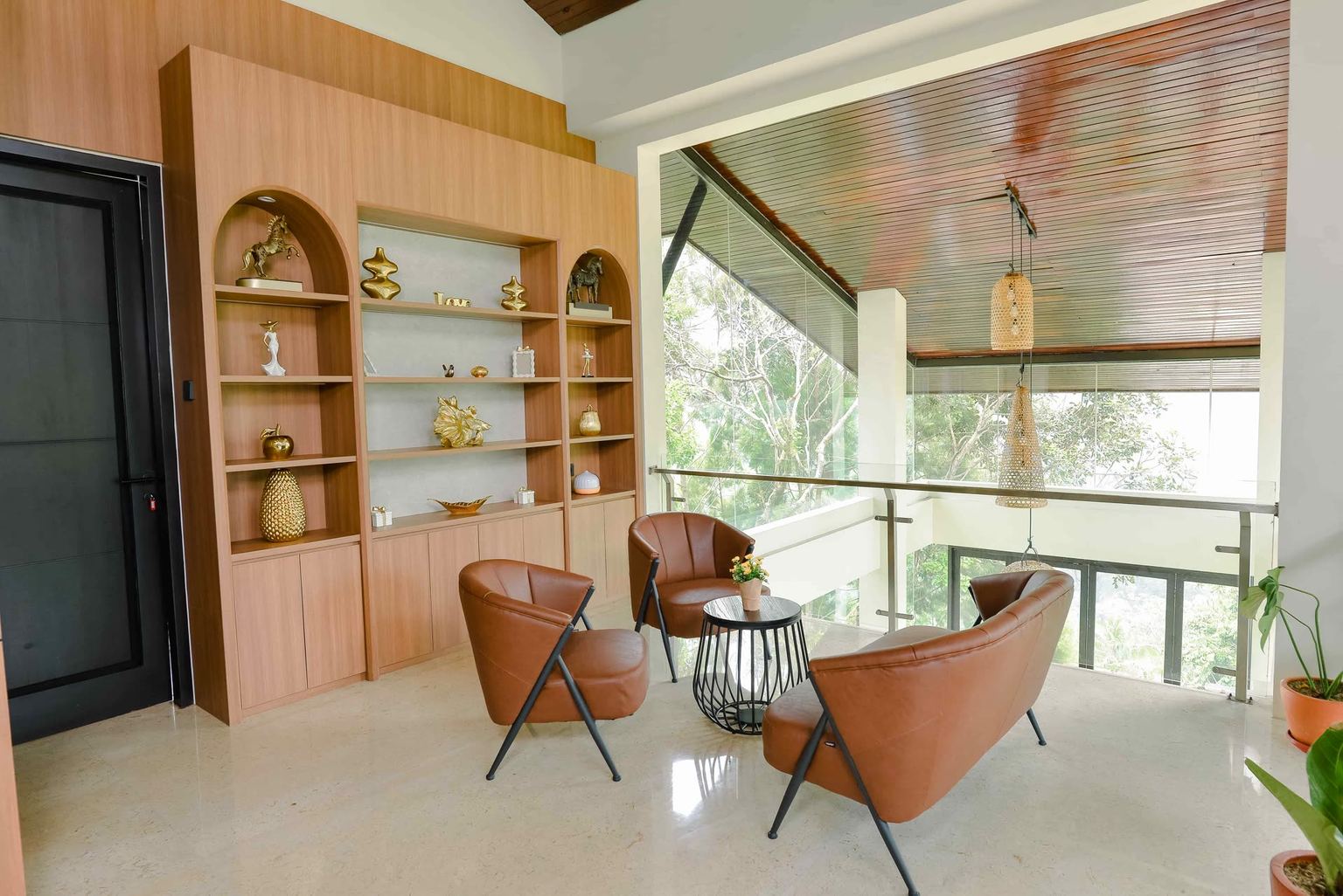 Lounge vila VANA SHANKARA dengan rak kayu, kursi kulit, dan jendela kaca menghadap pepohonan