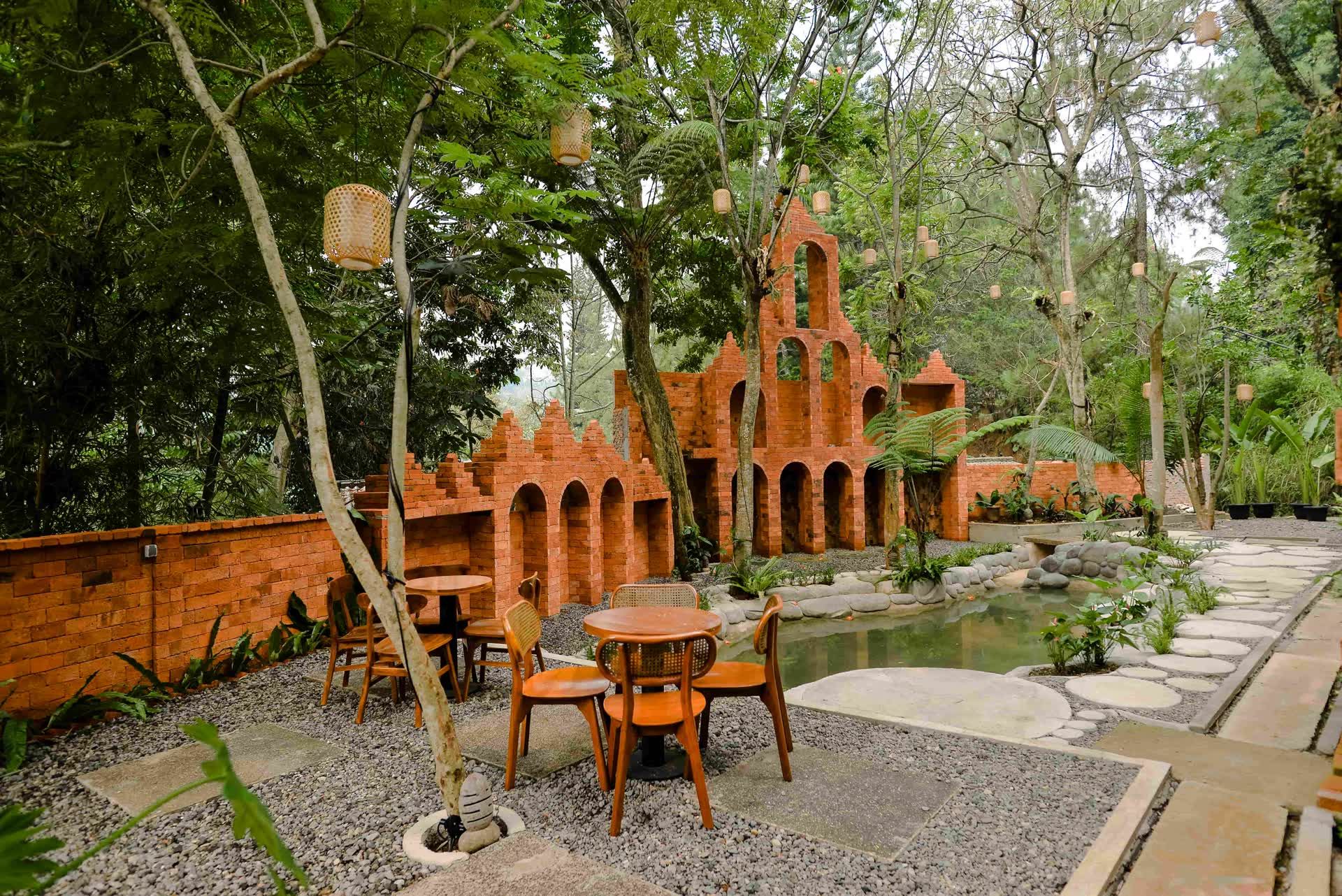 Meja dan kursi kayu di taman bata merah VANA SHANKARA dengan kolam dan pepohonan
