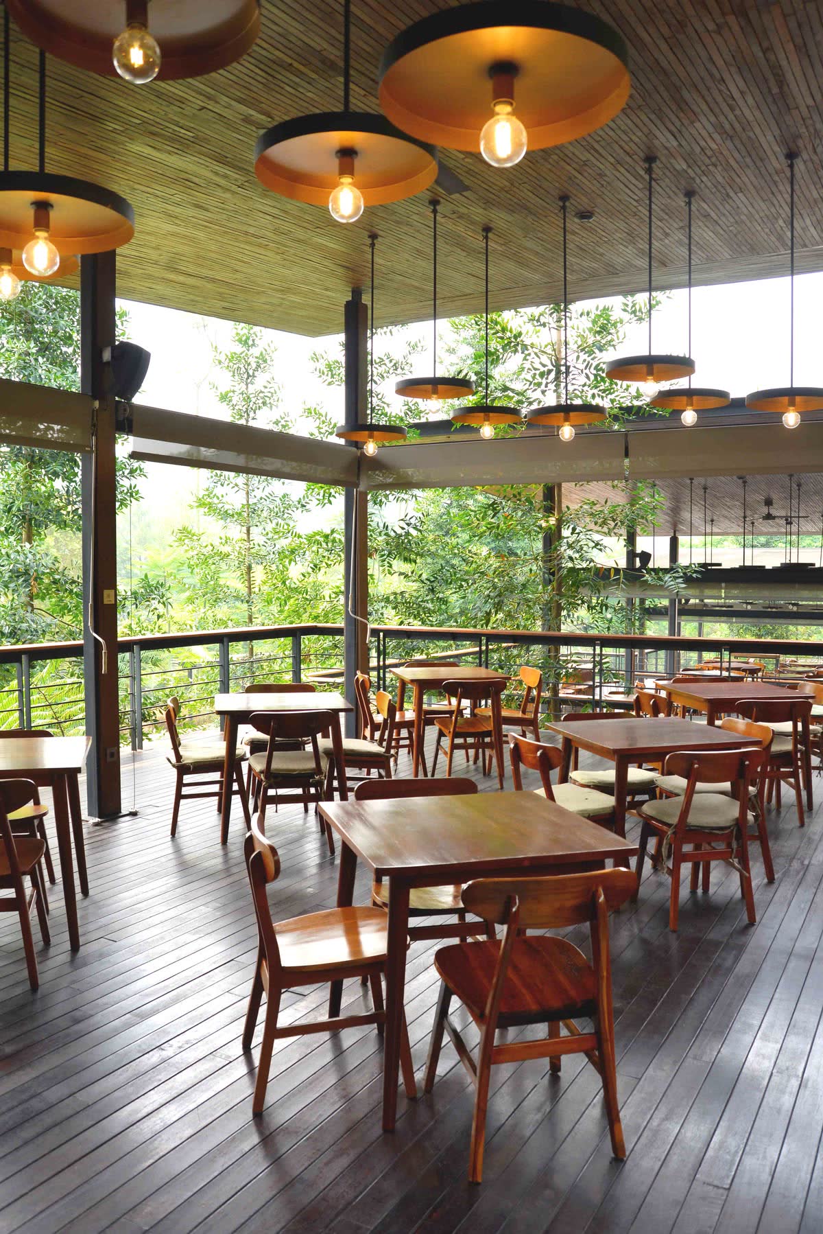 Meja kayu di teras resto VANA SHANKARA dengan lampu gantung dan pepohonan
